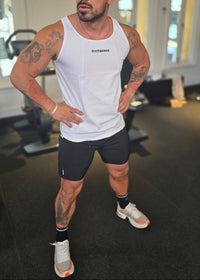 Man in a white tank top and black shorts standing indoors with a blurred background