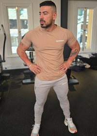 Man wearing a beige t-shirt and light gray pants standing indoors.