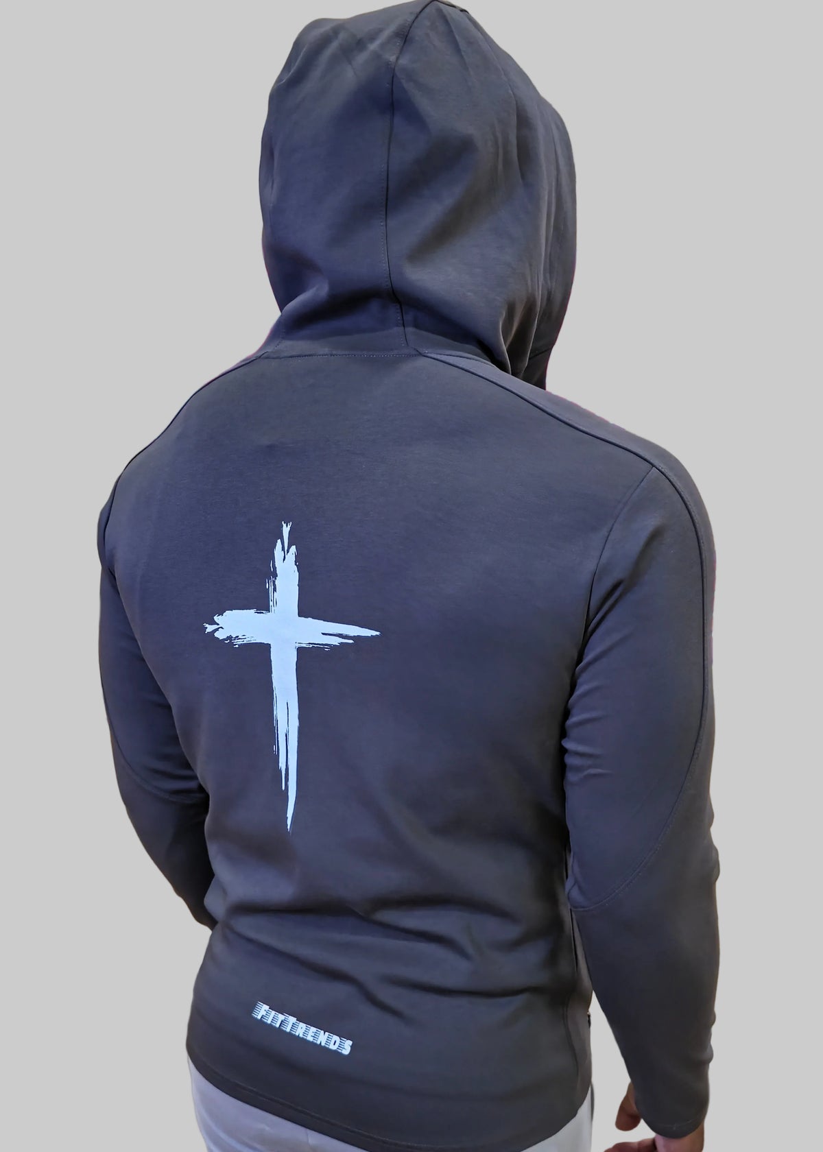 Person wearing a dark gray hoodie with a white cross design on the back, standing against a wooden panel wall.