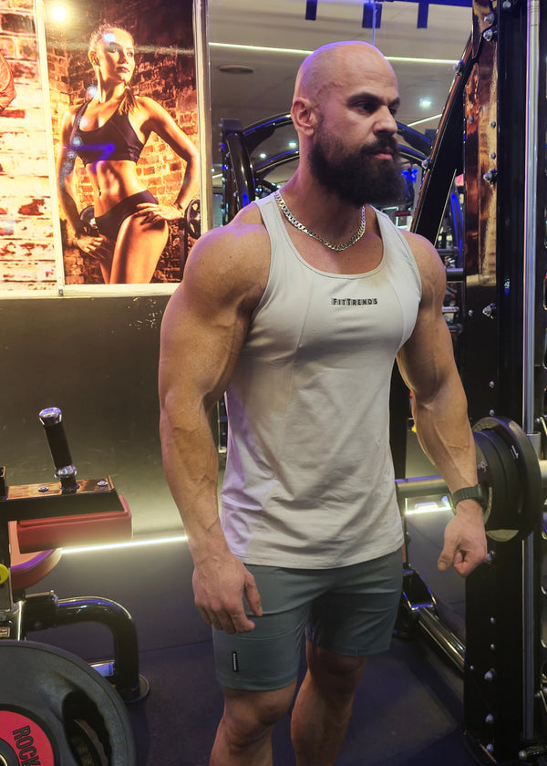 A muscular man with a beard stands in a gym wearing a white tank top with 'FITNESS' printed on the chest and gray shorts, surrounded by gym equipment and weight plates.