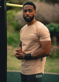Man wearing a beige t-shirt with a brand logo, standing outdoors.