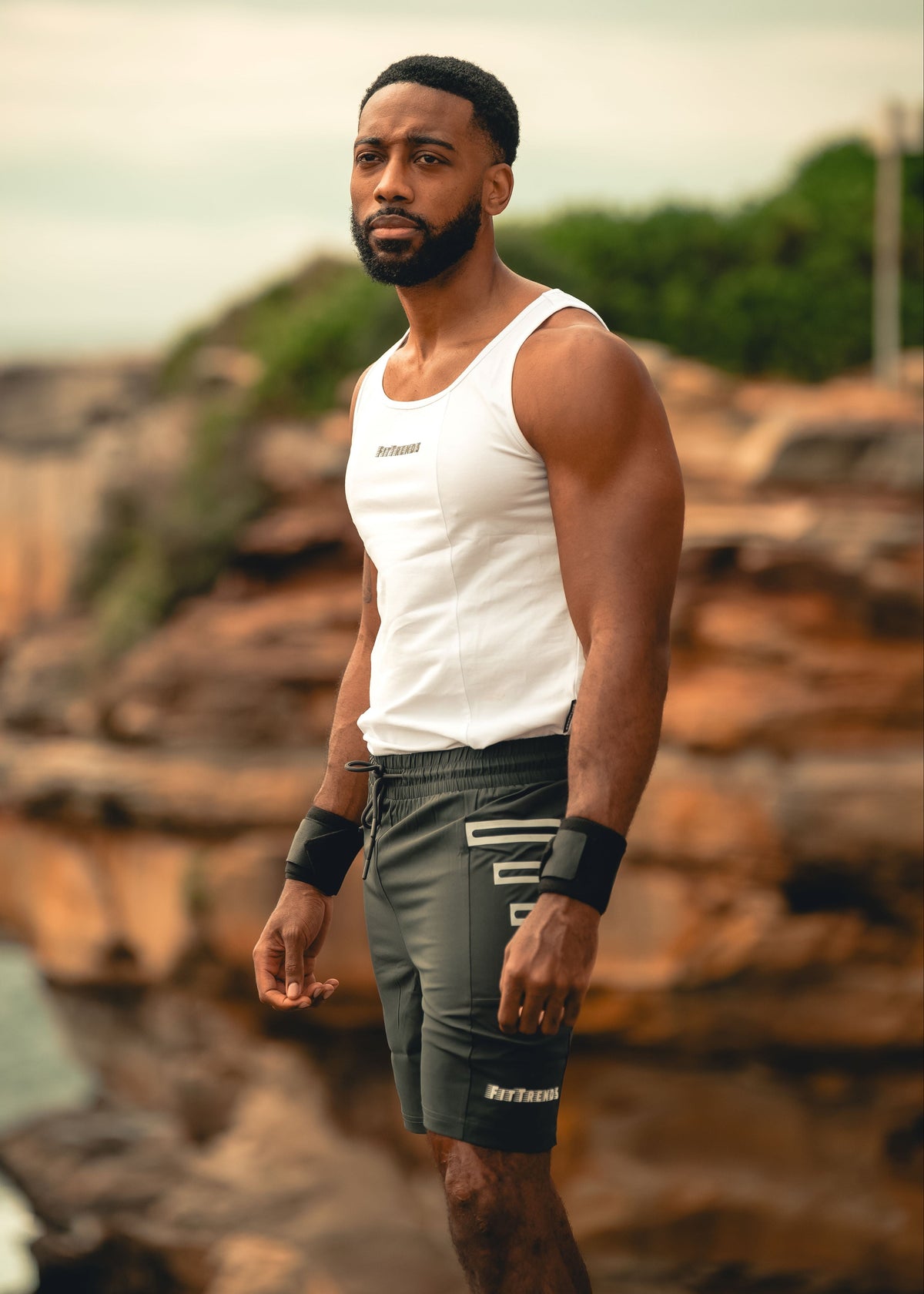 Man in a white tank top and gray shorts standing on a rocky shoreline with greenery in the background.