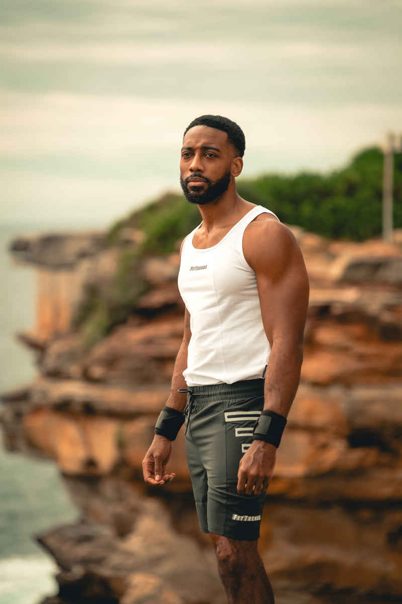 Man in a white tank top and gray shorts standing on a rocky shoreline with greenery in the background.