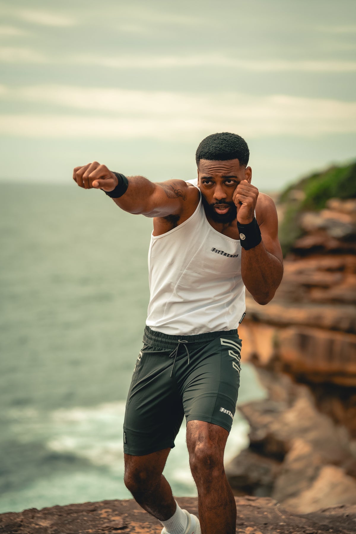 A man in athletic wear, featuring a white tank top and dark green shorts, posing in a fighting stance on a rocky cliffside overlooking the ocean.