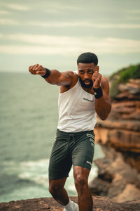A man in athletic wear, featuring a white tank top and dark green shorts, posing in a fighting stance on a rocky cliffside overlooking the ocean.