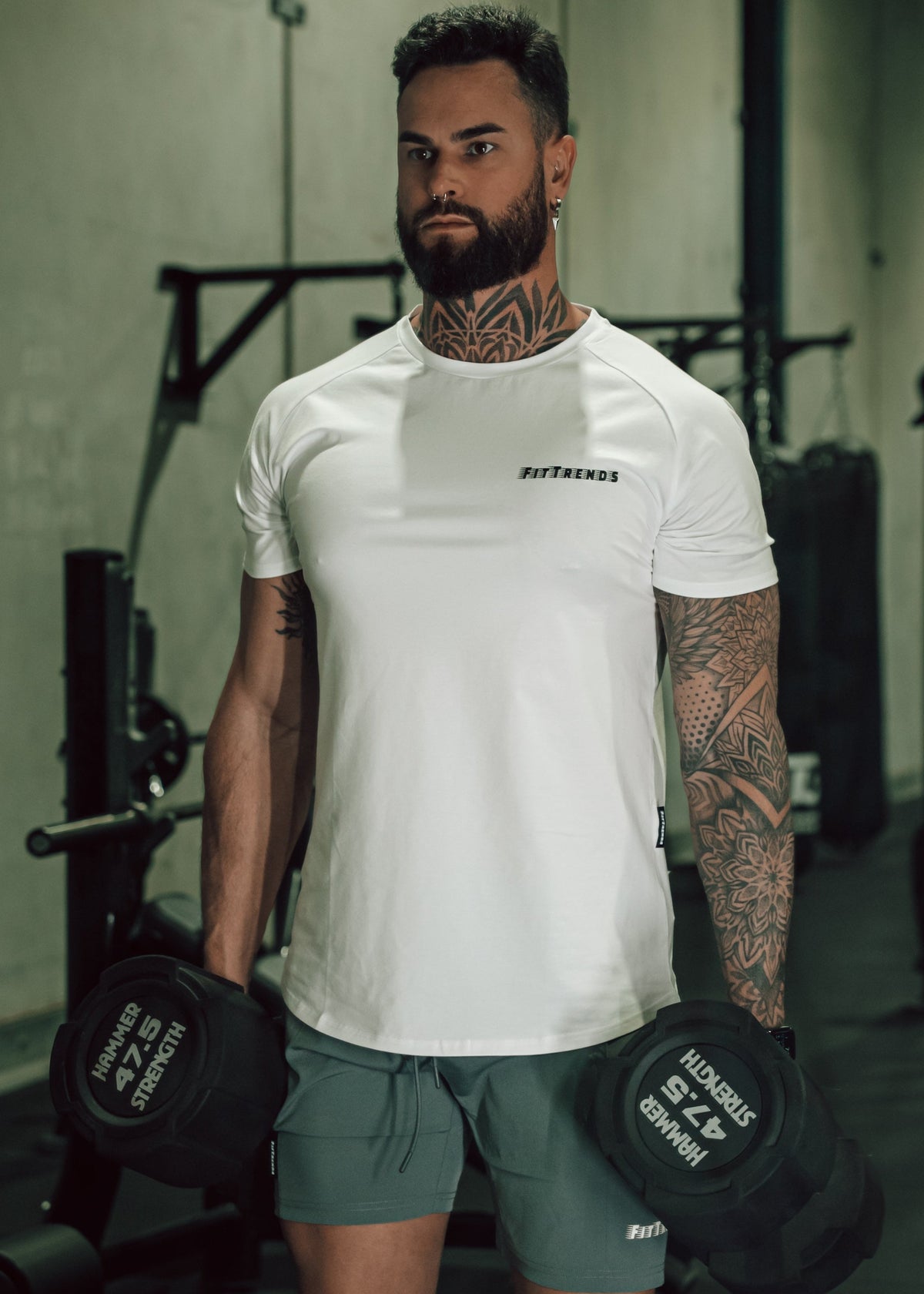 Man holding dumbbells in a gym wearing a white t-shirt with branding.