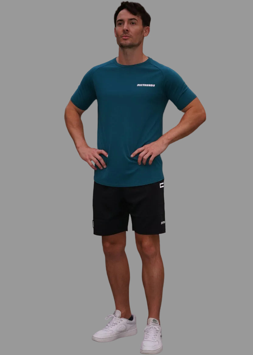 Man wearing a teal shirt and black shorts standing on a sidewalk with buildings and trees in the background.