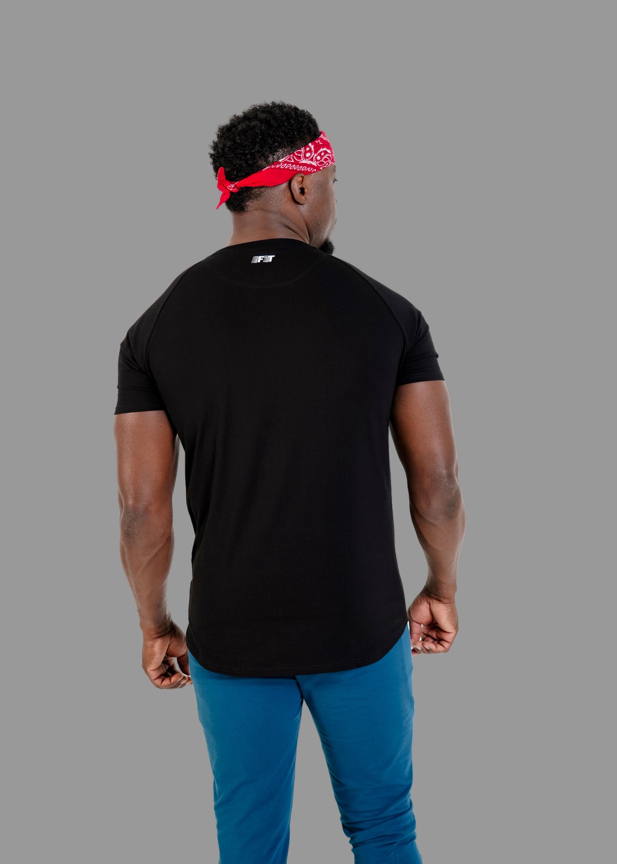 Person wearing a black t-shirt with a logo on the back, blue pants, and a red bandana, standing against a white background.