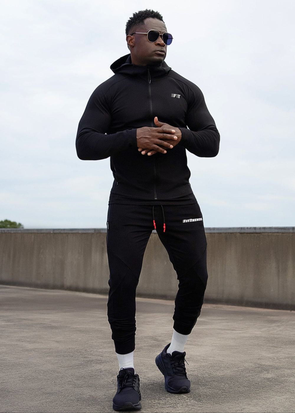 Man wearing a black tracksuit on a rooftop with a cloudy sky.