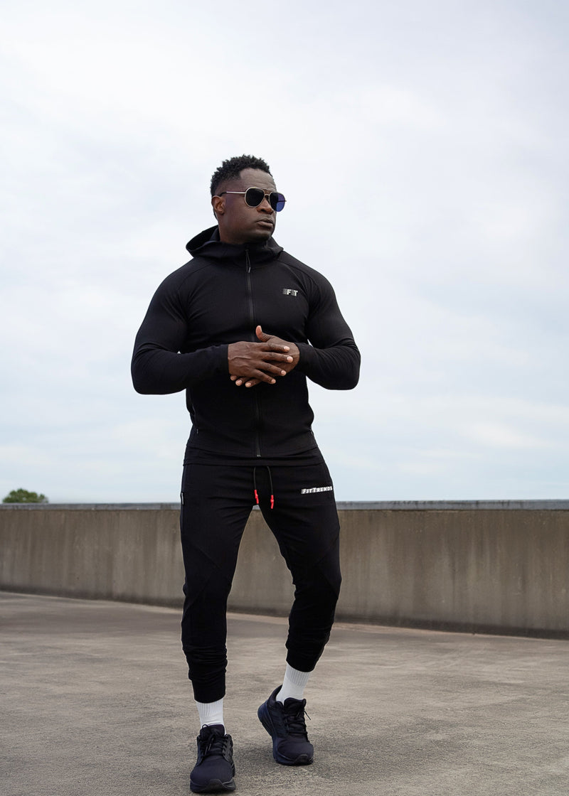 Man wearing a black tracksuit on a rooftop with a cloudy sky.