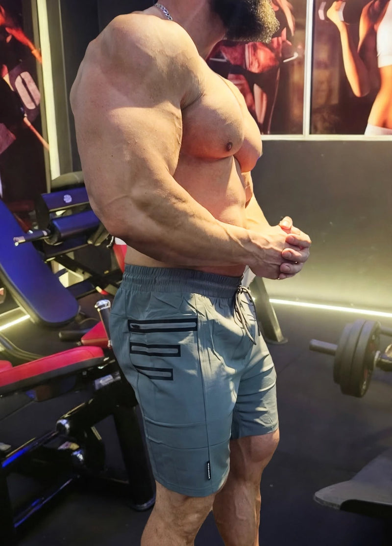 A muscular man in a gym wearing slate blue athletic shorts with black pocket details, posing in a fitness environment with weight machines in the background.