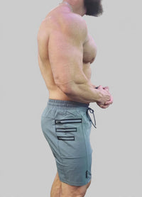 A side-profile view of a muscular man wearing slate blue-gray athletic shorts with black zipper details and a drawstring, set against a solid gray background.