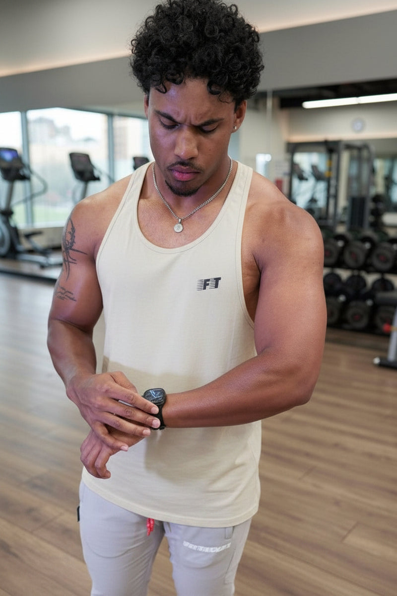 Man in a gym wearing a white tank top with a logo, looking at his wristwatch.