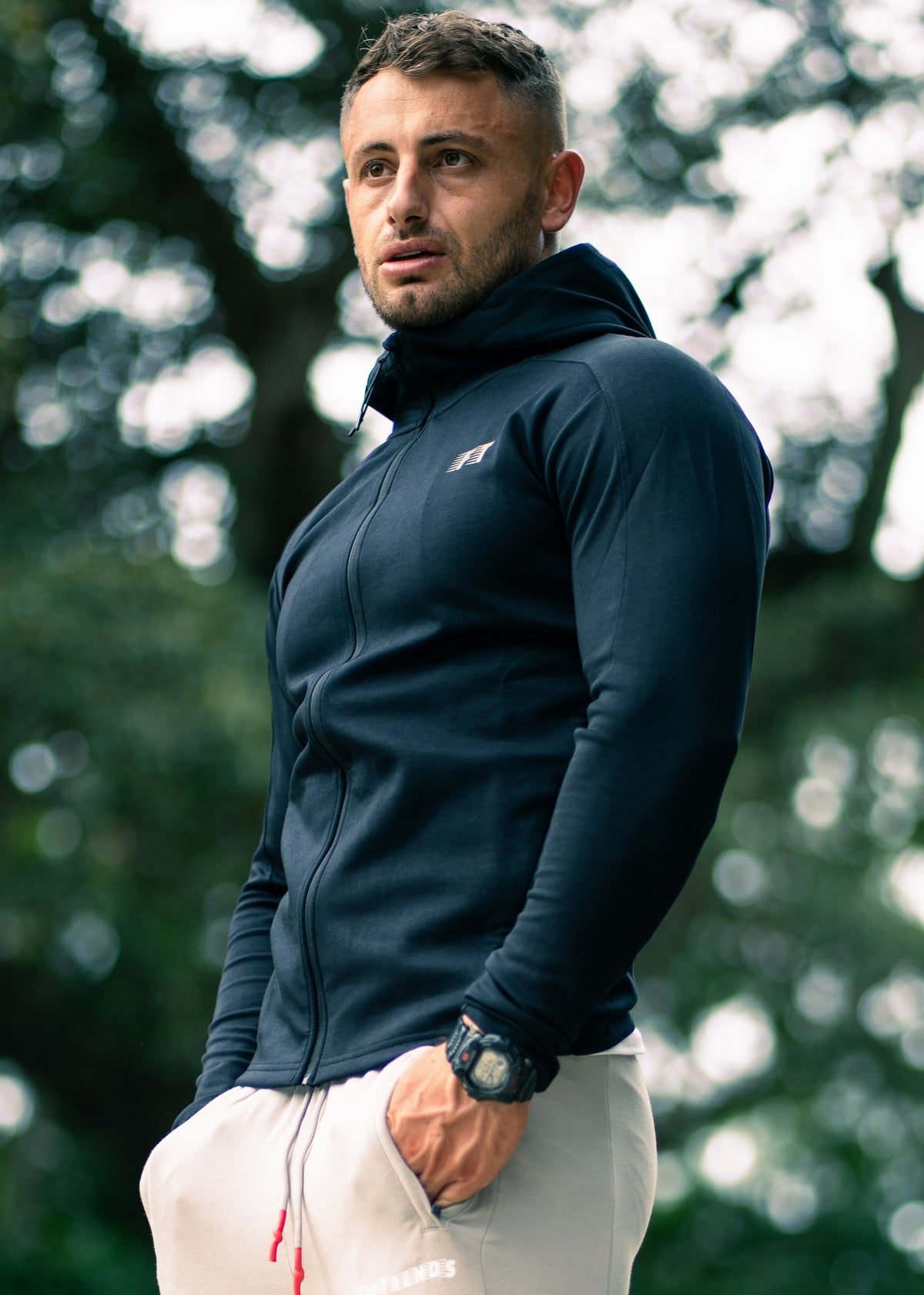 Man wearing a navy blue jacket with a brand logo, standing outdoors with blurred greenery in the background.