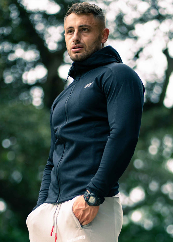 Man wearing a navy blue jacket with a brand logo, standing outdoors with blurred greenery in the background.